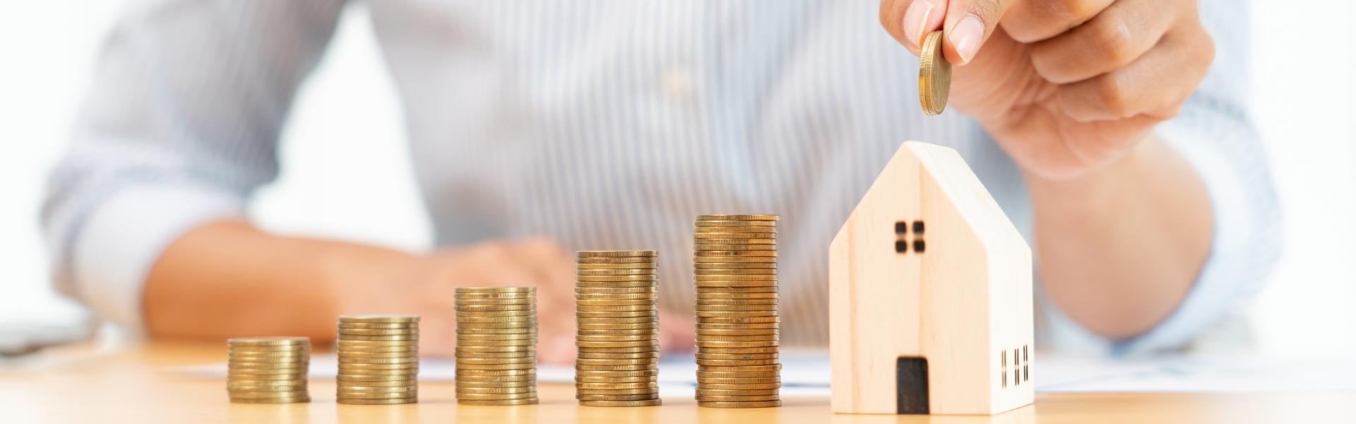 California VA Refinancing Options For Eligible Homeowners header image. Photo of a man putting a coin into a house-shaped piggybank. To the side of the house, are different stacks of coins making the shape of a bar graph to represent increase in value of the home.